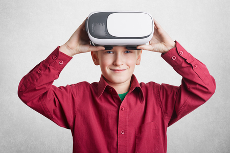 Handsome little male child wears VR glasses, has fun and entertains himself, plays video games, isolated over white background. Small boy experiences virtual reality, wears 3D glasses indoorの写真素材