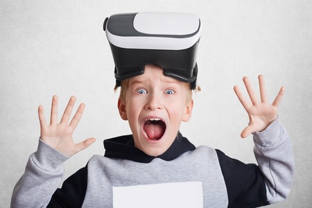 Excited small boy touches air during VR experience. Little child wears virtual reality headset on head, explores something, gestures actively. Entertainment and modern technologies conceptの写真素材