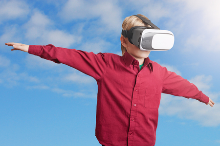 Freedom, technology and entertaiment concept. Small male child in red shirt wears VR glasses, studies possibilities of gadget, imagines that he flies in space, stands against clear sky backgroundの写真素材