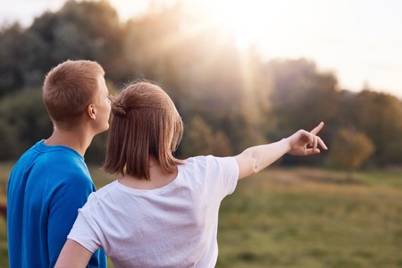 Back view of affectionate young couple stand closely, admire nature, see something into distance, enjoy sunset, copy space aside. Teenager indicates into distance, wants to show something beautifulの写真素材