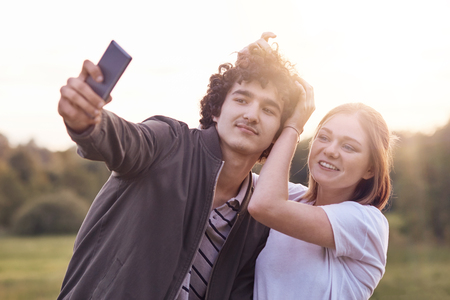 Funny curly male youngster and his girlfriend pose for making selfie portrait against blurred nature background, have positive expressions. Couple in love use modern cell phone for entertainmentの写真素材