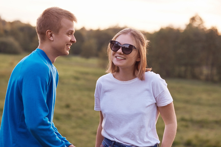 Happy romantic couple spend free time outdoor, have fun together, foolish, pose against meadow background. Joyful teenage girl in shades, casual white t shirt looks positively at her boyfriendの写真素材