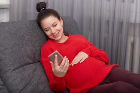 Pregnancy, leisure, technology concept. Happy smiling pregnant girl holds smart phone, wears red...