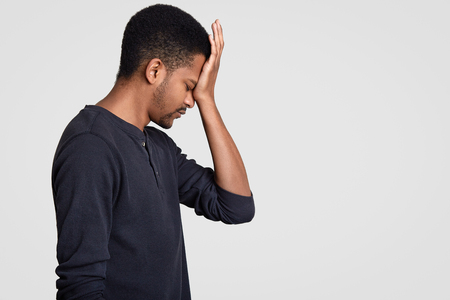 Profile shot of dark skinned man in casual outfit, hits his forehead with regret, being pessimist, feels unhappy, being in despair, stands against white wall with free space for your advertisementの写真素材