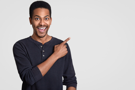 Positive dark skinned man with toothy smile, points at upper right corner with index finger, suggests use copy space, wears casual clothing, has funny expression, stands against white studio wallの写真素材