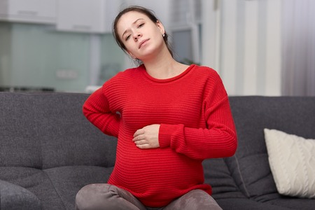 Photo of tired displeased pregnant woman feels backache, keeps hand on swollen belly, sits at couch...