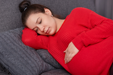 Photo of restful young woman with dark hair combed in bun, keeps hand on swollen tummy, dressed in knitted loose sweater, lies on sofa, anticipates for child. Feminity and fertility concept.の写真素材