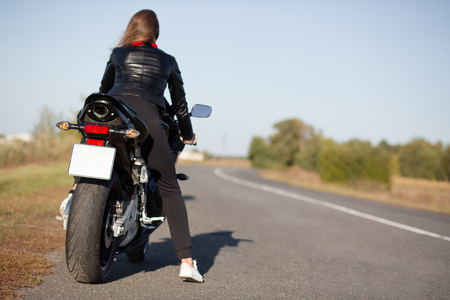 Back view of fast female motorcyclist wears leather jacket, poses on motorbike, takes break after racing competitions, free space for your advertising content over road background. Adrelnaline conceptの写真素材
