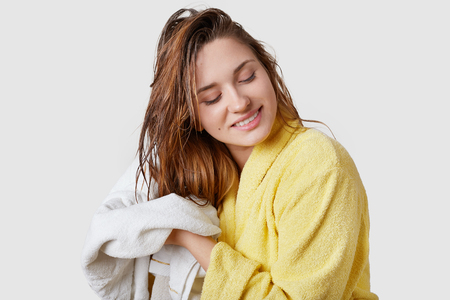 Indoor shot of beautiful woman wipes wet hair with towel, dressed in yellow bathrobe, stands against white background, has pleased facial expression, has shower at home, satisfied with new shampooの写真素材