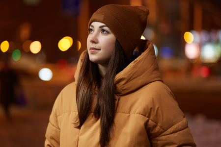 Outdoor shot of thoughtful young woman wears stylish hat and brown jacket, has pensive facial expression, enjoys calm atmosphere in evening city, poses over blurred background. Lifestyle conceptの写真素材
