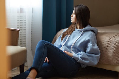 Indoor shot of thoughtful dark haired woman dressed in blue sweatshirt, jeans, sits on floor near bed, looks thoughtfully at window, poses in cozy bedroom at modern apartment. Domestic atmosphereの写真素材