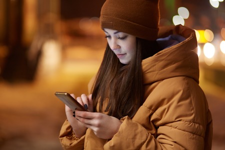 Cropped image of pleasant looking woman concentrated in smart phone, dressed in stylish headgear and jacket, has outdoor walk in evening, searches information in internet. Blurred city view.の写真素材