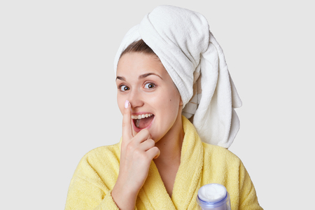 Funny attractive young woman keeps fore finger on nose, applies beauty cream, stands in bathroom, wears white towel and yellow robe, stands against white studio wall. Cosmetic and women conceptの写真素材