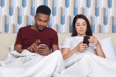 Domestic atmopshere, technology and ethnicity concept. Concentrated mixed race young woman and man use modern mobile phones, pose at bed in cozy hotel, have lazy day, chat in social networksの写真素材