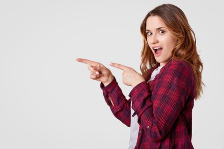 Horizontal shot of cheerful young woman with long hair, points with both fore fingers at empty space, wears casual checkered shirt, shows direction to somewhere, isolated over white background.の写真素材