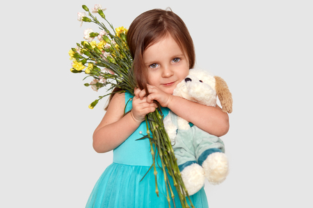 Studio shot of attractive child carries tightly flowers and soft toy, dressed in puffy blue dress, isolated over white background, recieves present from father on 8 March. Childhood concept.の写真素材