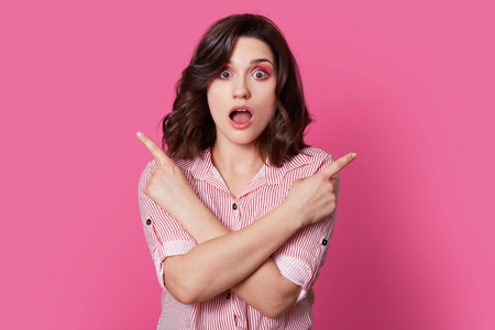 Photo of surprised young European woman with make up, crosses hands over chest, opens mouth, dressed in striped shirt, isolated over pink background. People, advertisement, reaction concept.の写真素材