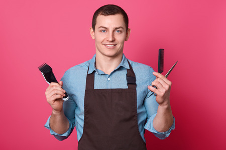 Positive friendly looking barber with electric razor, scissors and comb, dressed in shirt and apron, ready to do hairstyle for men, poses in babershop against pink background. Grooming conceptの写真素材