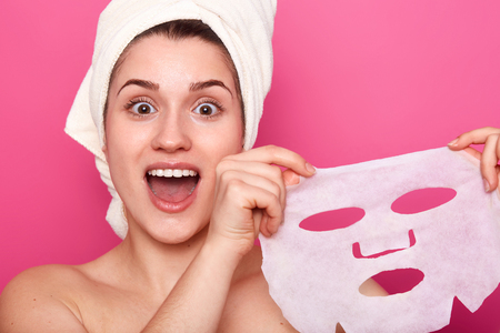 Headshot of amazed beautiful young woman holds cosmetic mask, feels refreshed and energized, has wrapped towel on head, looks with amazement directly at camera, isolated over pink backgroundの写真素材