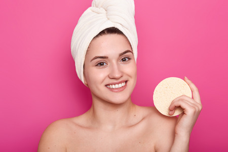 Smiling European woman with cheerful expression, shows white teeth, holds sponge, looks direclty at camera, enjoys softness after taking shower, models over pink background. Body care conceptの写真素材
