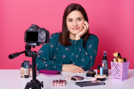 Pleased female famous blogger records video for her beauty blog, advertises different cosmetics products, wears fashionable blouse, sits at white desk against pink background. Beauty industryの写真素材