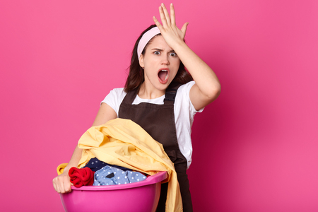 Forgetful female keeps hand on forehead, remembers she forgot to buy washing powder, dressed in white casual t shirt and apron, carries basin with pile of clothes, isolated on pink backgroundの写真素材