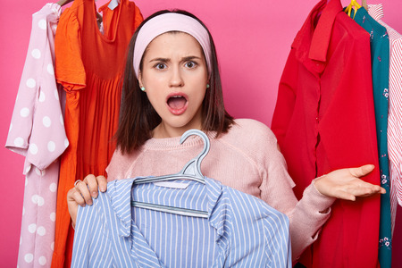 Surprised brunette woman with opened mouth, raises palm. Dark haired girl holds hanger with blue striped blouse. Lady dislikes so high price for shirt. Female wears sweater and hair band in showroom.の写真素材