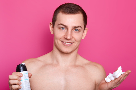 Positive blue eyed man carries bottle of shaving gel, poses with naked torso, showsc bare shoulders, muscles, has gentle smile, has smooth sking after shaving, isolated over pink background.の写真素材