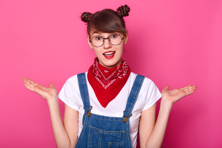 Young brunette student in confusion, stand with open mouth isolated over pink background. Schoolgirl with two funny bunches, wears casual white t shirt and denim overalls, makes helpless gesture.の写真素材