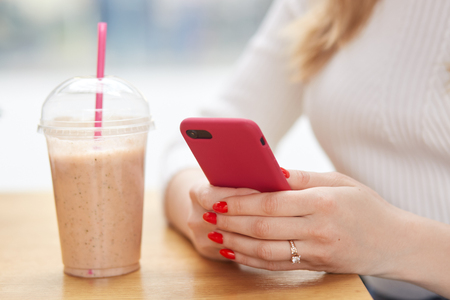 Faceless woman holds modern cell phone in hands, has red manicure, checks notification, drinks milkshake from plastic cup with straw, connected to wireless internet in cafeteria. Technology conceptの写真素材