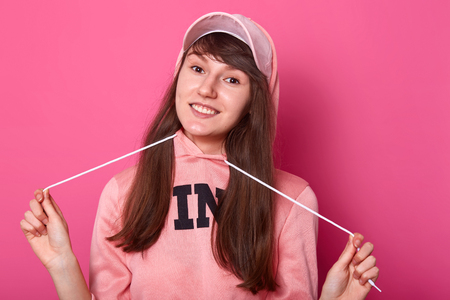 Beautiful charming girl wears pink hoodie and cap. Young lady has dark long hair, fantastic smile and brown eyes. Tenager with hood on head stands isolated over rose background. Teens concept.の写真素材