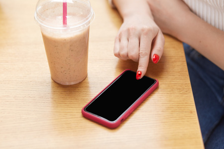 Unrecognizable woman uses modern cell phone, keeps finger with red nail on touchscreen, drinks milk cocktail, mock up screen for your advertisement or promotional content. People, leisure, technologyの写真素材