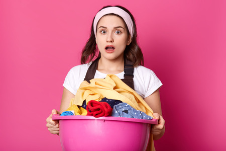 Young woman holds basin full of clean linen, housewife looks tired after doing laundry, works about house, dresses brown apron, white t shirt and hair band, stands with open mouth. Copy space.の写真素材