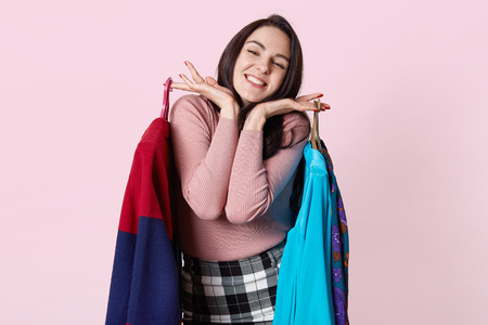 Portrait of beautiful happy brunnette female with dark hair, poses against pink wall, being in good...