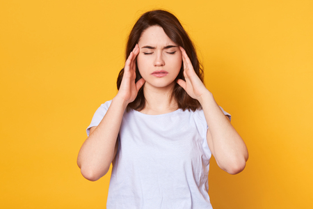 Image of young good looking brunette female stands with closed eyes isolated over yellow background, keeps hands on temples, has awful headache. Charming girl wears white t shirt. Healthy concept.の写真素材