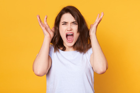 Image of cute young lady standing isolated over yellow background, looking directly at camera, screaming something, keeps hands near head, wears white casual t shirt. Copy space for advertisment.の写真素材