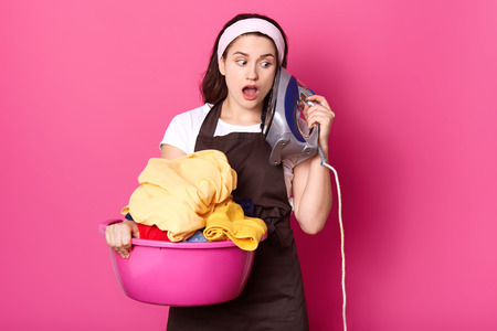 Close op portrait of cute good looking young woman with surprised facial expression, holds pink basin full of clean clothes and keeps iron like phone, imagine conversation, has fun while ironig.の写真素材