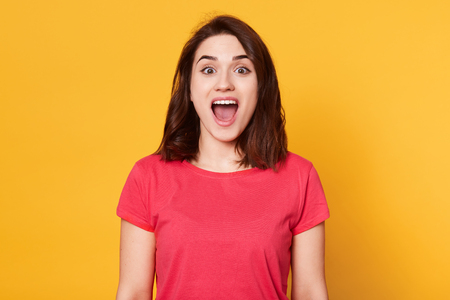 Surprised young woman shouting over yellow background, looking at camera, looks happy, has wonderful news, being in good mood, glad to hear something pleasant, posing in studio. People emotion conceptの写真素材