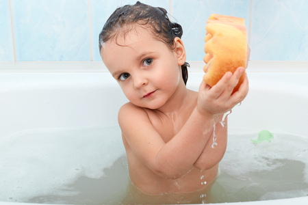 Beautiful infant sitting around foam in bathroom puts washcloth in two hands, trying to squeeze it, focused on washing process. Attractive little kid is curious about taking bath. Care concept.の写真素材