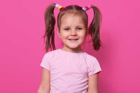 Close up portrait of cheerful sweet little girl with funny pigtails, smiling sincerely, looking directly at camera, standing straight, having colourful scrunchies. Copy space for advertisement.の写真素材