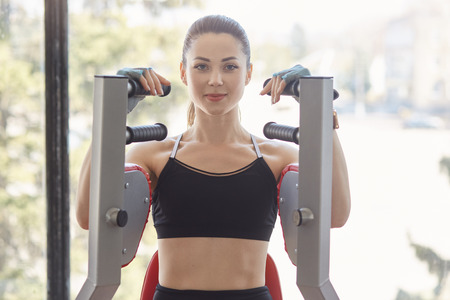 Blue eyed strong European woman spends her free time in gym, does work sets, looks directly at camera, seems to be pleased. Charming model poses at training simulator, sitting in front of window.の写真素材