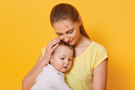Close up portrait of beautiful happy mother hugging her baby girl with love, mom wears casual t shit with ponytail, pose in photo studio isolated over yellow background. Motherhood and family concept.の写真素材