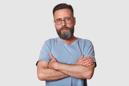Close up portrait of handsome man with beard standing with arms folded isolated on white background, looks serious, wearing casual gray t shirt and glasses, male posing in photo studio. People conceptの写真素材