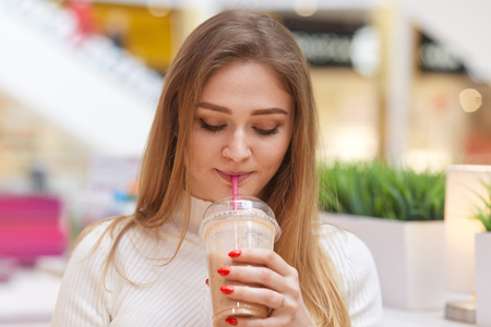 Photo of charming blonde female drinks cocktail, poses in cafe, wears white casual sweater, has red manicure, looks down at her beverage, likes its taste, enjoying going out, People and youth concept.の写真素材