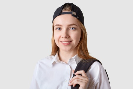 Smiling positive school girl looks directly at camera, poses isolated over white background, holding black bag pack with one hand, wearing white shirt and black cap, looks delighted. Study concept.の写真素材