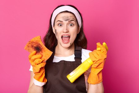 Impressed tired model posing isolated over pink background in studio, opening mouth with shock, doing household chores, wearing brown apron, t shirt and white headband. Housewife and cleaning concept.の写真素材