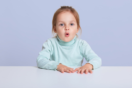 Image of astonished lovely little sitting at white desk, small female child with rounded lips looking with surprised expression.の写真素材