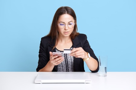 Portrait of concentrated professional business woman using mobile phone and credit card at her work, sitting at white desk, making online payment, female wearing elegant black outfit and glasses.の写真素材