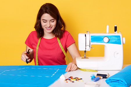 Image of brunette girl wears red t shirt, sitting isolated over yellow background, holds scissors in hand, has charming smile, sews stylish dress, sitting surrounded with different sewing equipment.の写真素材