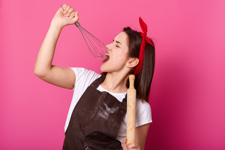 Studio shot of young baker wears kitchen apron smeared with flour, white t shirt, red headband, holds baking rolling pin, whisk like microphone, stands singing isolated over pink background.の写真素材